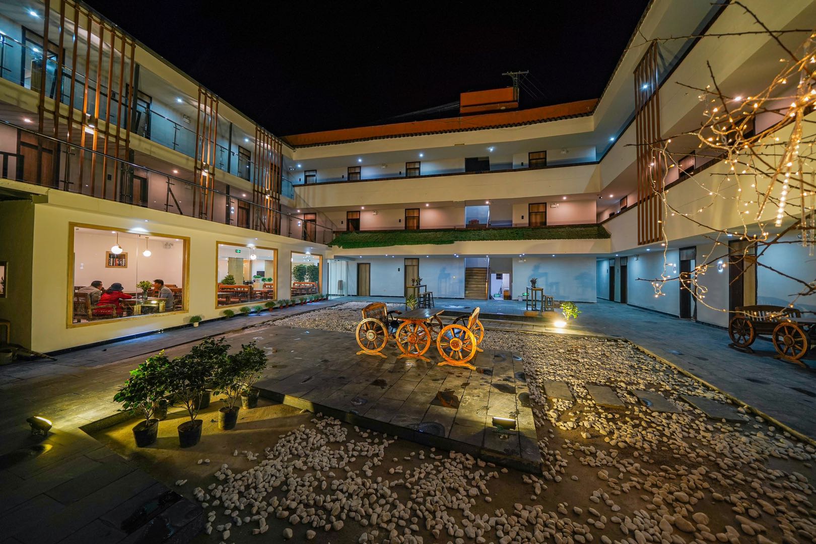 夜里的内院：三层围合的客房阳台亮着灯，地上是石子与木台，树枝上缠着暖色灯串